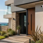 Modern house entry with a wooden door, stone facade, and a landscaped path toward a coastal view.