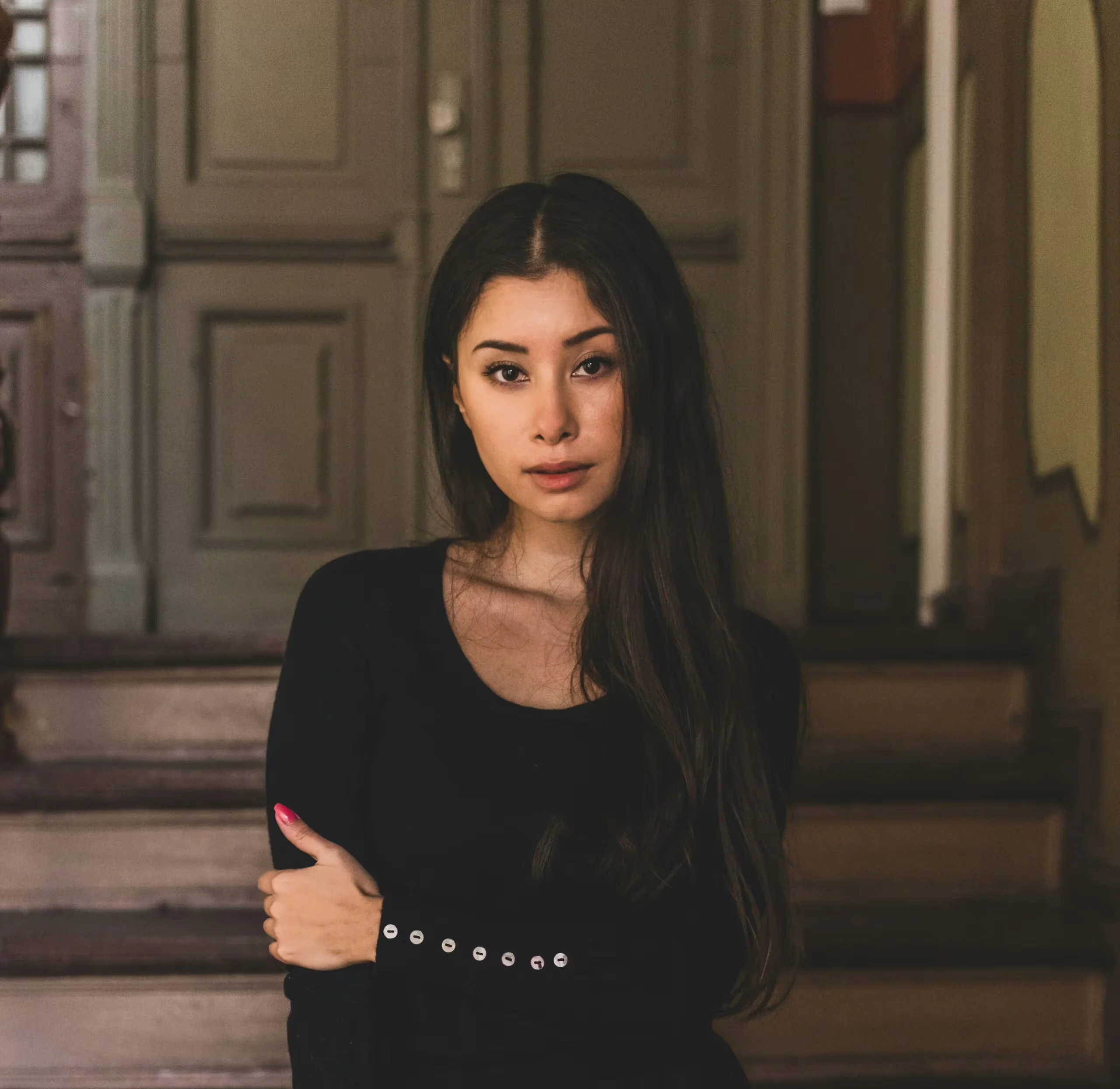 Portrait of a woman with long dark hair, wearing a black top, standing with arms crossed in a doorway.