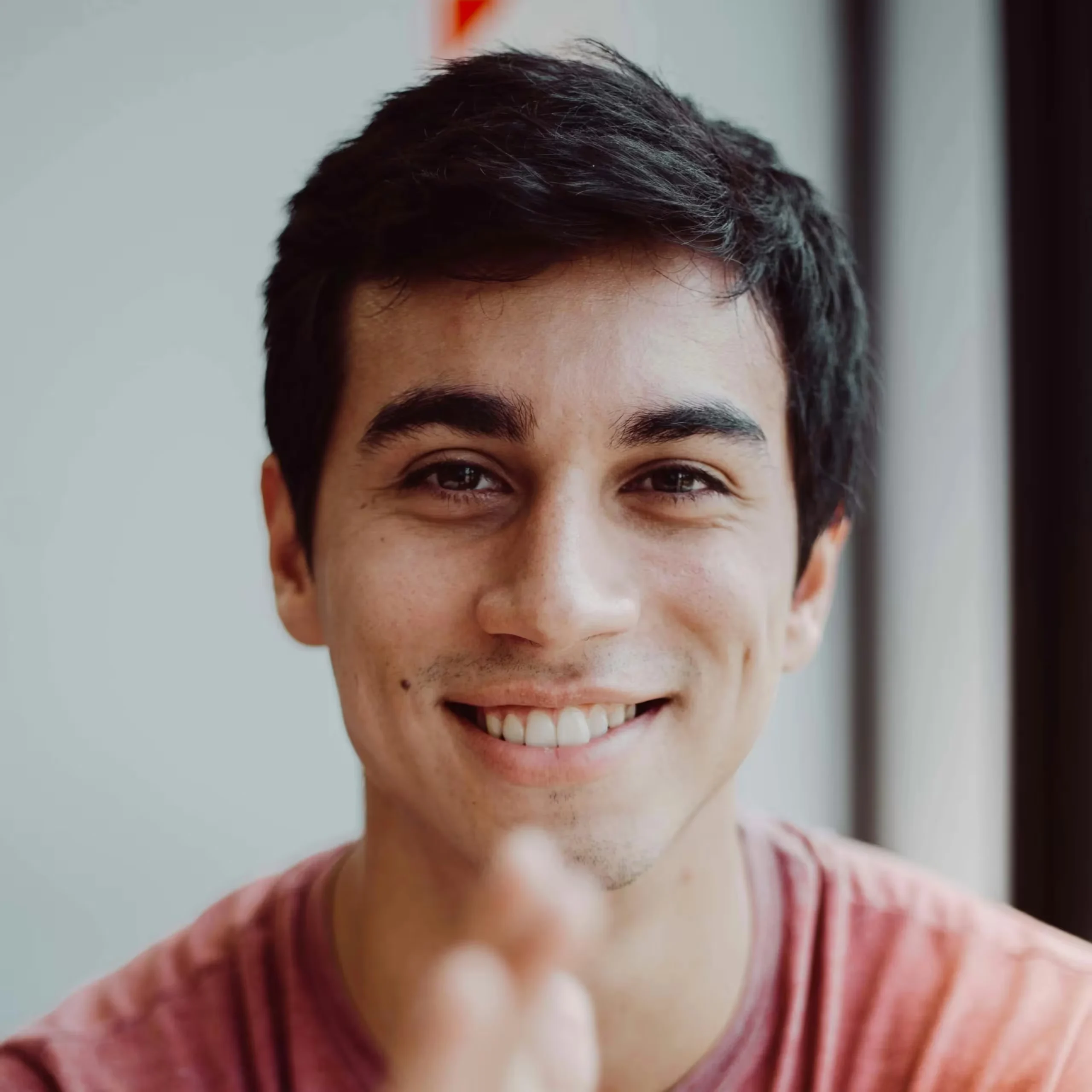 Close-up of a smiling man with dark hair, wearing a pink striped shirt, against a neutral background.