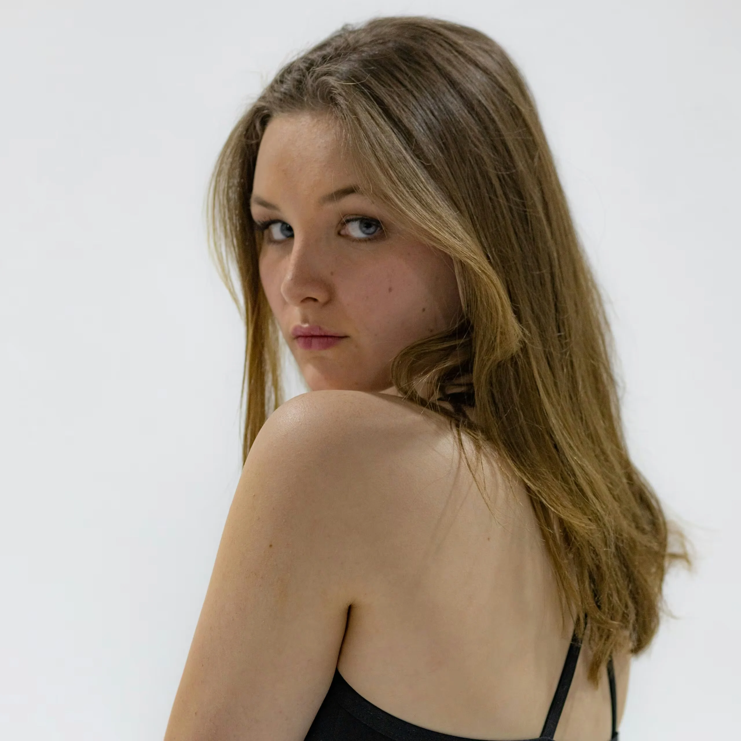 Young woman with long brown hair looking over her shoulder, wearing a black spaghetti-strap top against a light background.