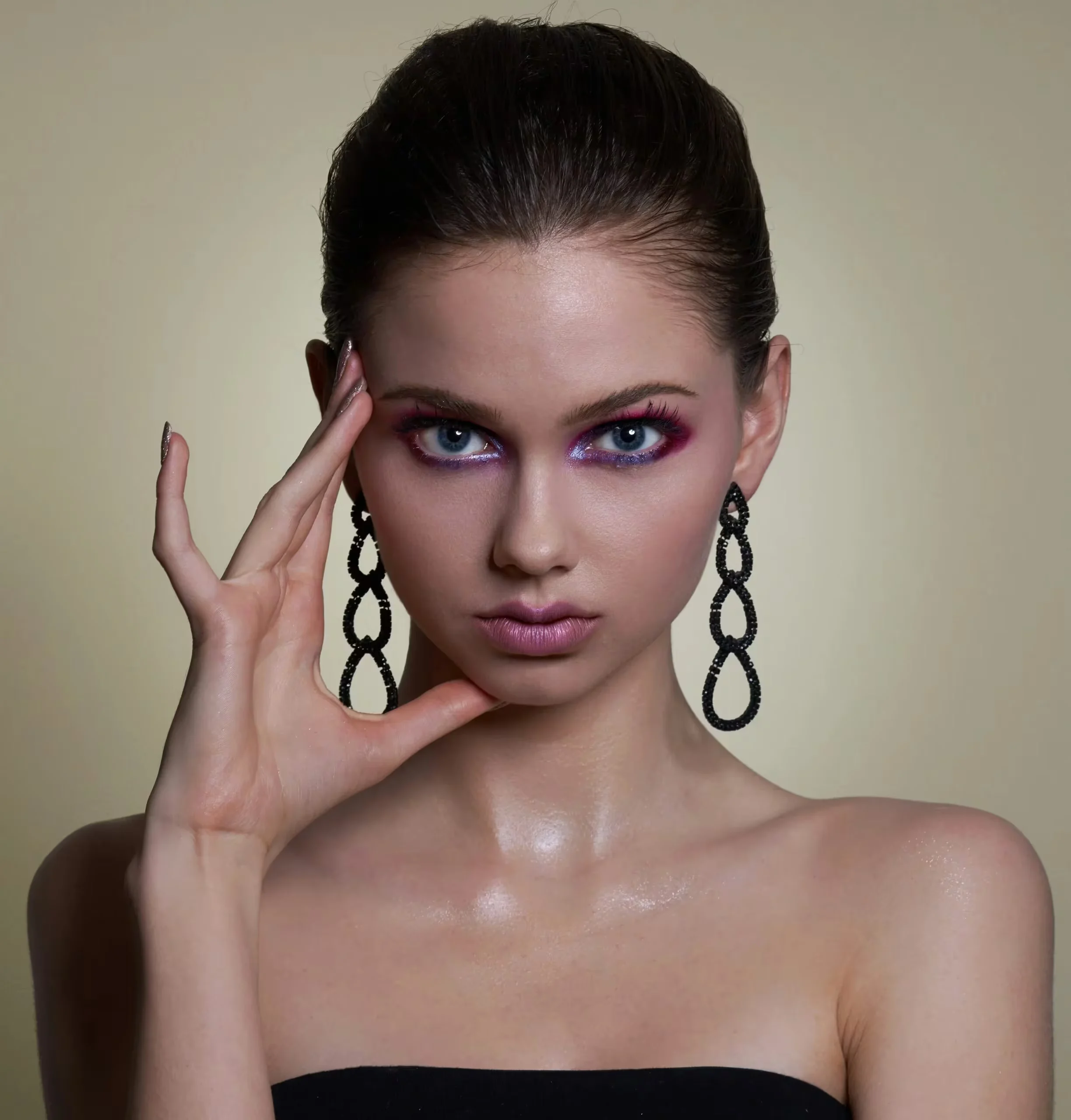 Close-up of a woman with dramatic pink and purple eye makeup, hair slicked back, wearing large black teardrop earrings.